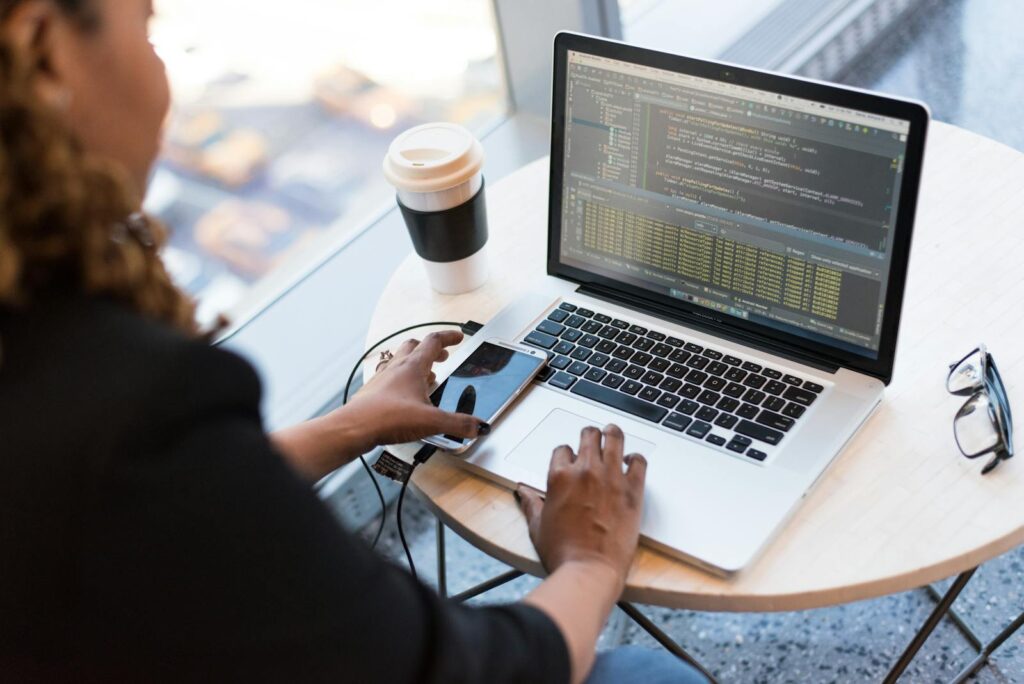 The Best Custom Web Development Solutions for Your Business Needs 16 Black woman programming on a laptop with coffee, smartphone, and glasses on a desk in an office.