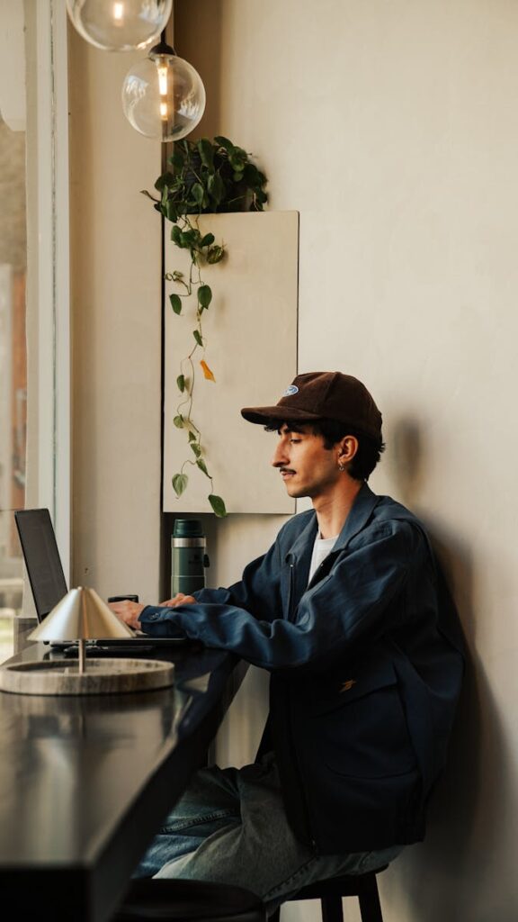 Young man wearing cap and jacket works on laptop in a stylish cafe with warm lighting and green plants.