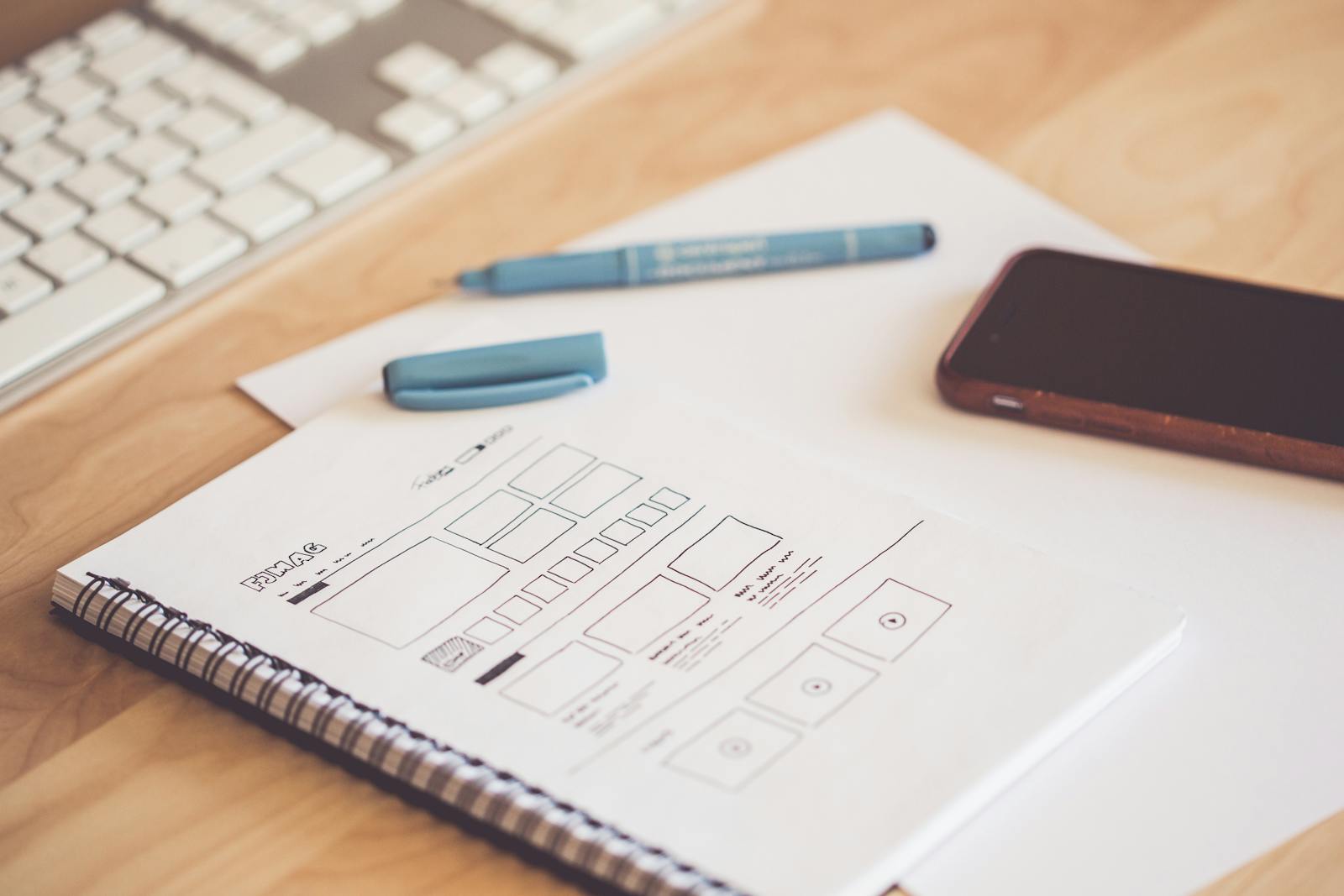 A neat workspace featuring a sketchpad with wireframes, a smartphone, and a keyboard on a wooden desk. (Small Business Web Design) | Building Sites That Convert & Grow Your Brand